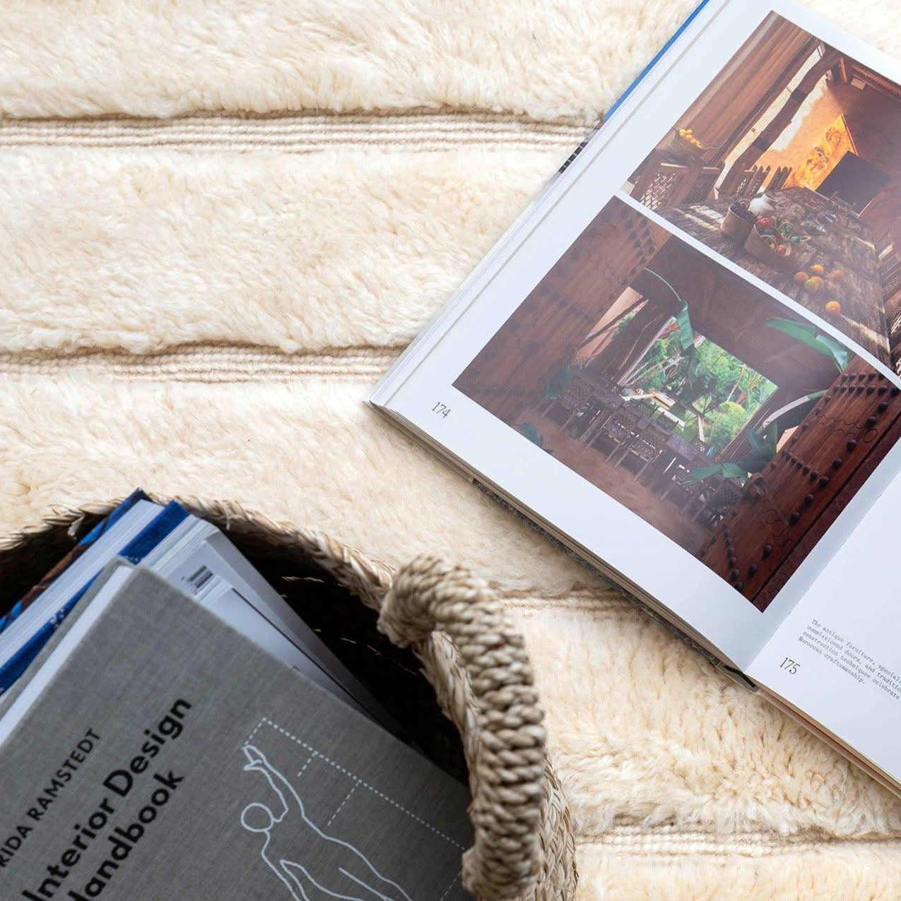 An open book with vivid interior design photos placed on a soft, cream-colored wool rug from Barbary Wool, alongside a woven basket filled with design handbooks.