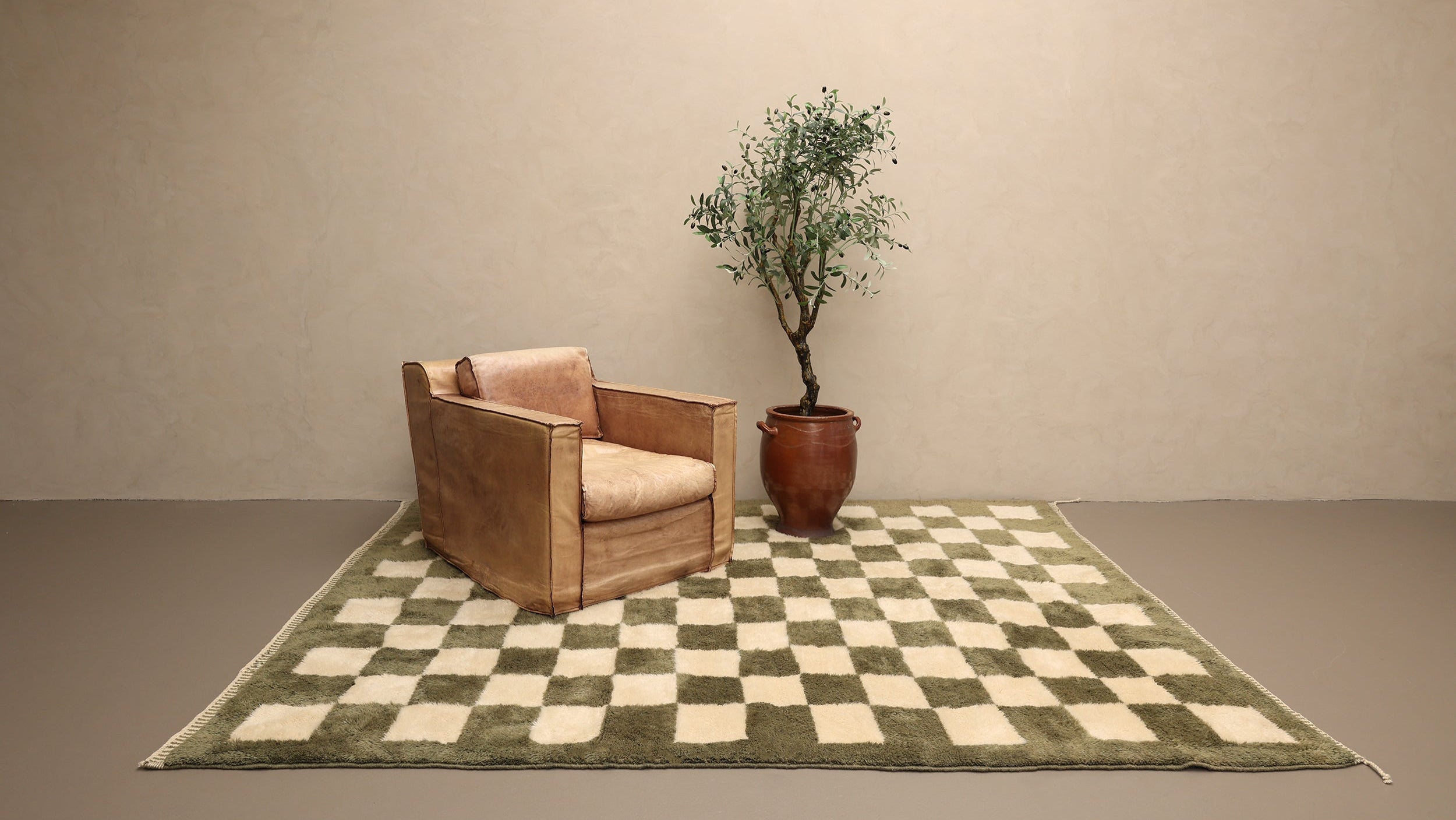 Leather armchair on a checkered forest green Berber rug with an olive plant against a beige wall.