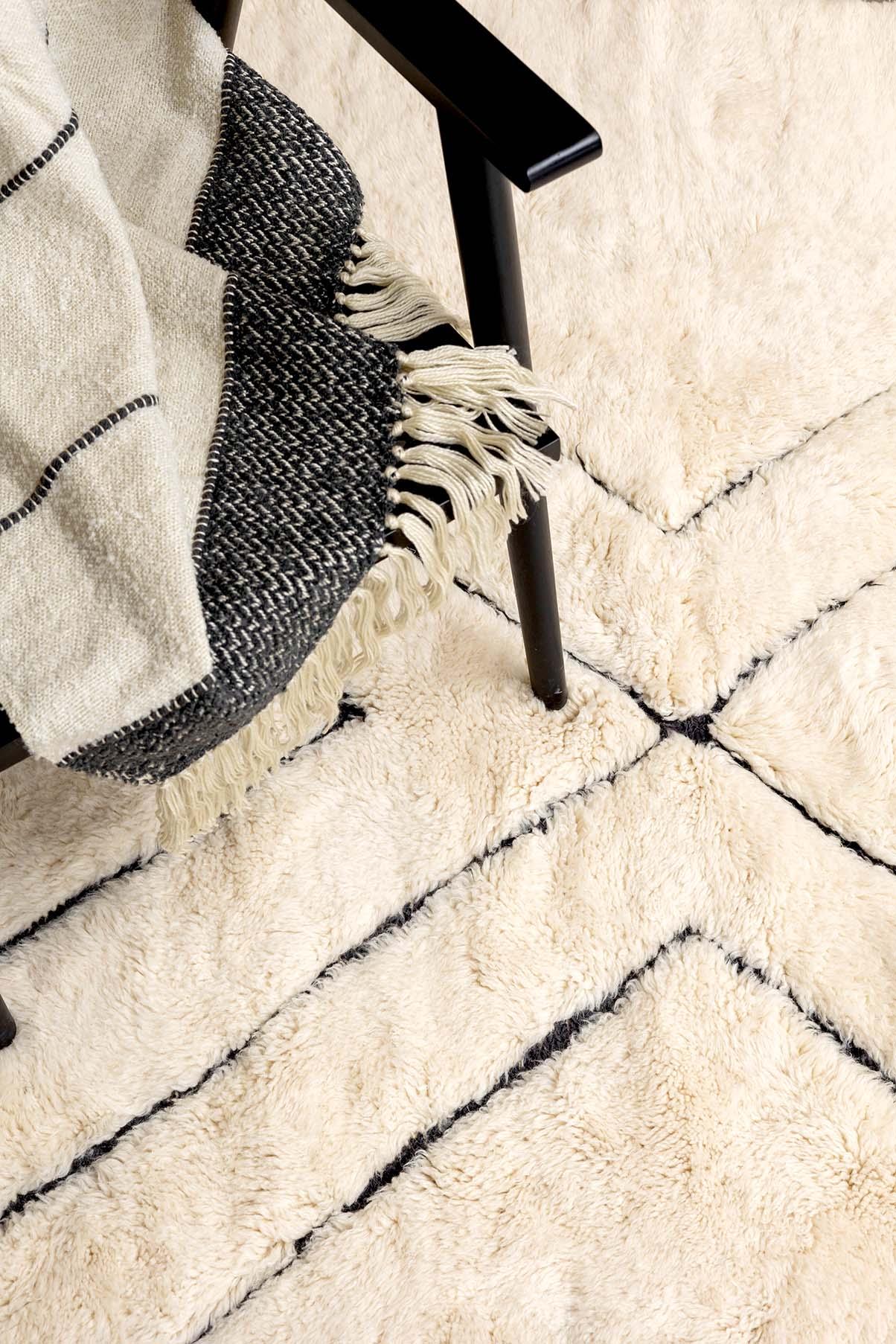 Close-up view of Barbary Wool's Azrou rug showcasing its cozy, textured surface and minimalist chevron design, complemented by a soft woven throw and a sleek chair leg.