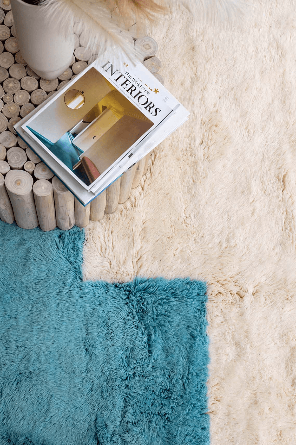 Vibrant composition showcasing Barbary Wool's NASNAS Collection rug in a dual-tone design—contrasting cream and teal textures—accentuated by a rustic cylindrical table and a contemporary interiors magazine, creating a cozy yet modern aesthetic.