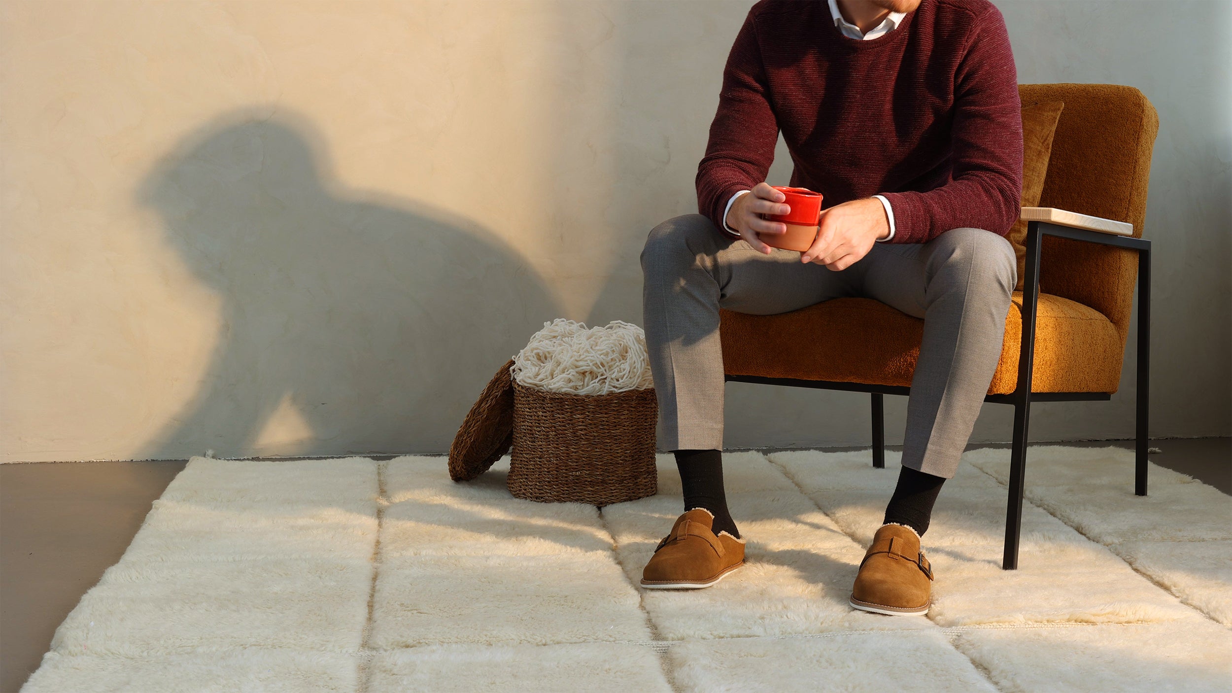 Man sitting on a mustard-colored armchair, holding a red cup, with a woven basket and white throw placed nearby on a soft beige barbary wool rug, creating a cozy and minimalist indoor ambiance.